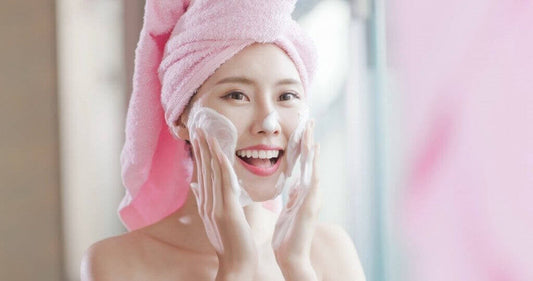 Smiling woman with a pink towel applying facial cleanser for healthy skin care routine.