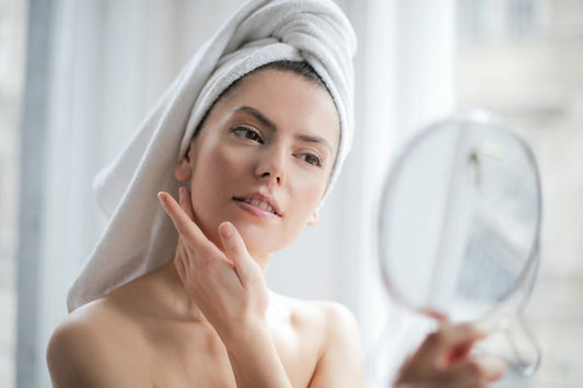 Woman in a towel examining her skin in a mirror, focusing on skincare routine for a healthy glow.