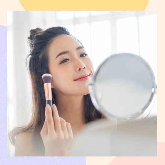 Woman applying makeup with a brush while looking in the mirror, showcasing a radiant K-beauty glow.