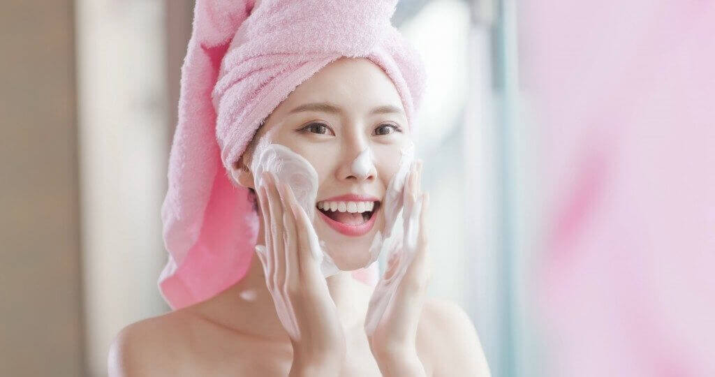 Smiling woman with a pink towel applying facial cleanser for healthy skin care routine.