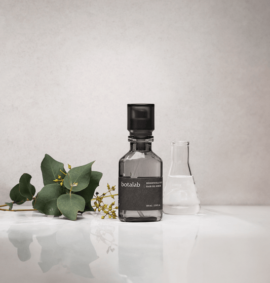 Botalab Deserticola Plus Hair Oil Serum bottle on a white surface with greenery, showcase of hair care product.